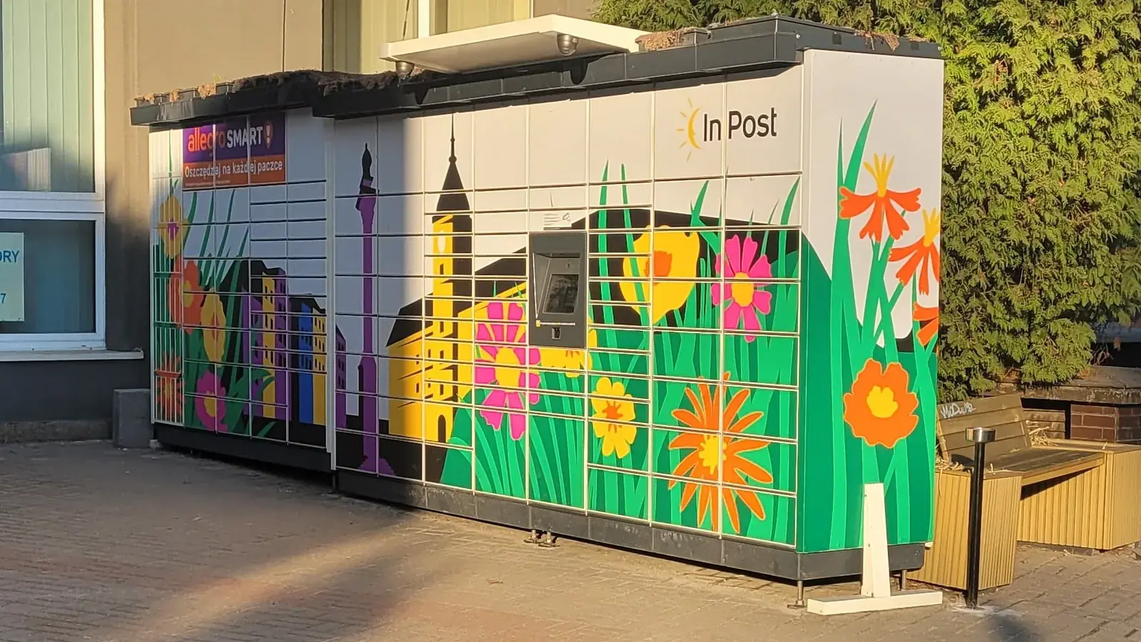 InPost przekierowanie paczki do Paczkomatu - automat paczkowy InPost przy ulicy Mikołaja Kopernika w Warszawie, obudowa z malowanymi kwiatami i sylwetką Kolumny Zygmunta, w popołudniowym słońcu na chodniku z kostki